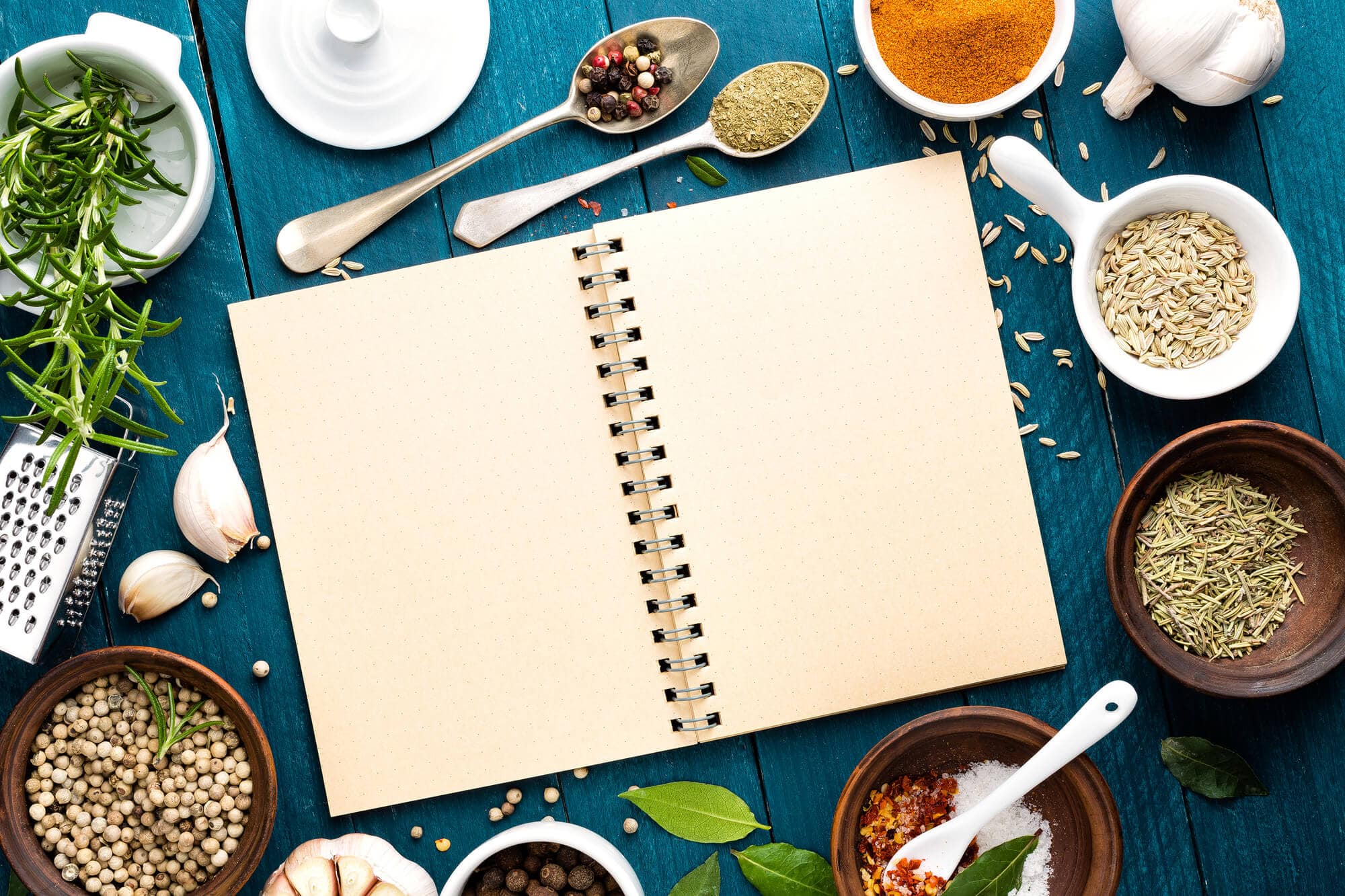 Empty recipe book against a teal wooden background featuring cooking utensils, dishware, herbs and spices