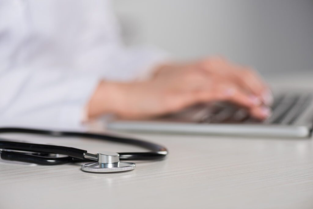 A physician typing on a laptop with a stethoscope in front and in focus