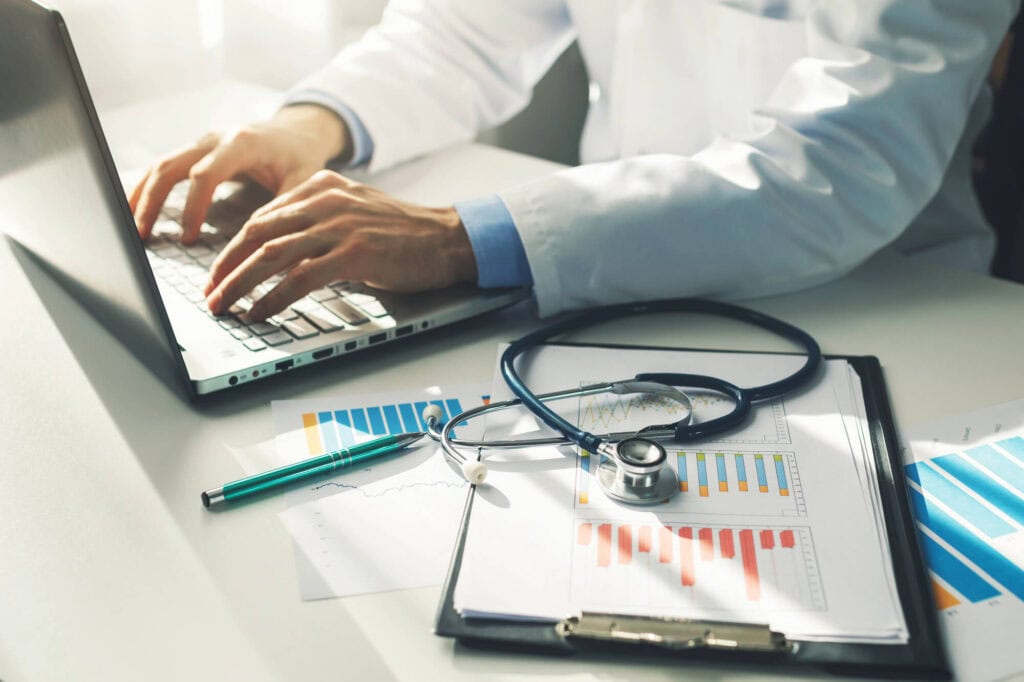A healthcare provider in a white coat types on a laptop, surrounded by a stethoscope, pen, and charts displaying colorful data on a clipboard. This scene highlights the integration of hospital automation systems.