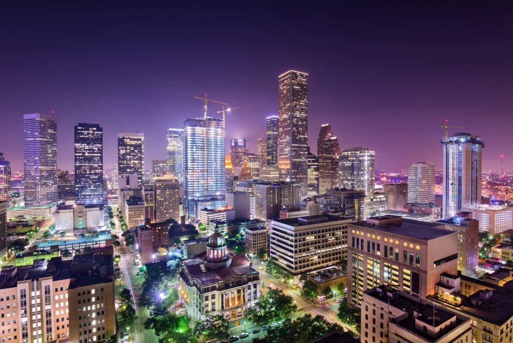 A vibrant city skyline at night with tall, illuminated skyscrapers, construction cranes, and a historic courthouse surrounded by city buildings. The sky glows purple, and streets below are lit with city lights.