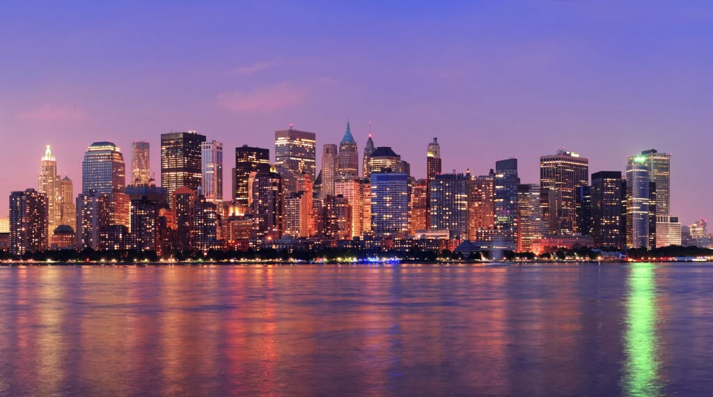 A city skyline at dusk with tall buildings lit up, their lights reflecting on the calm water in the foreground under a purple and pink sky.