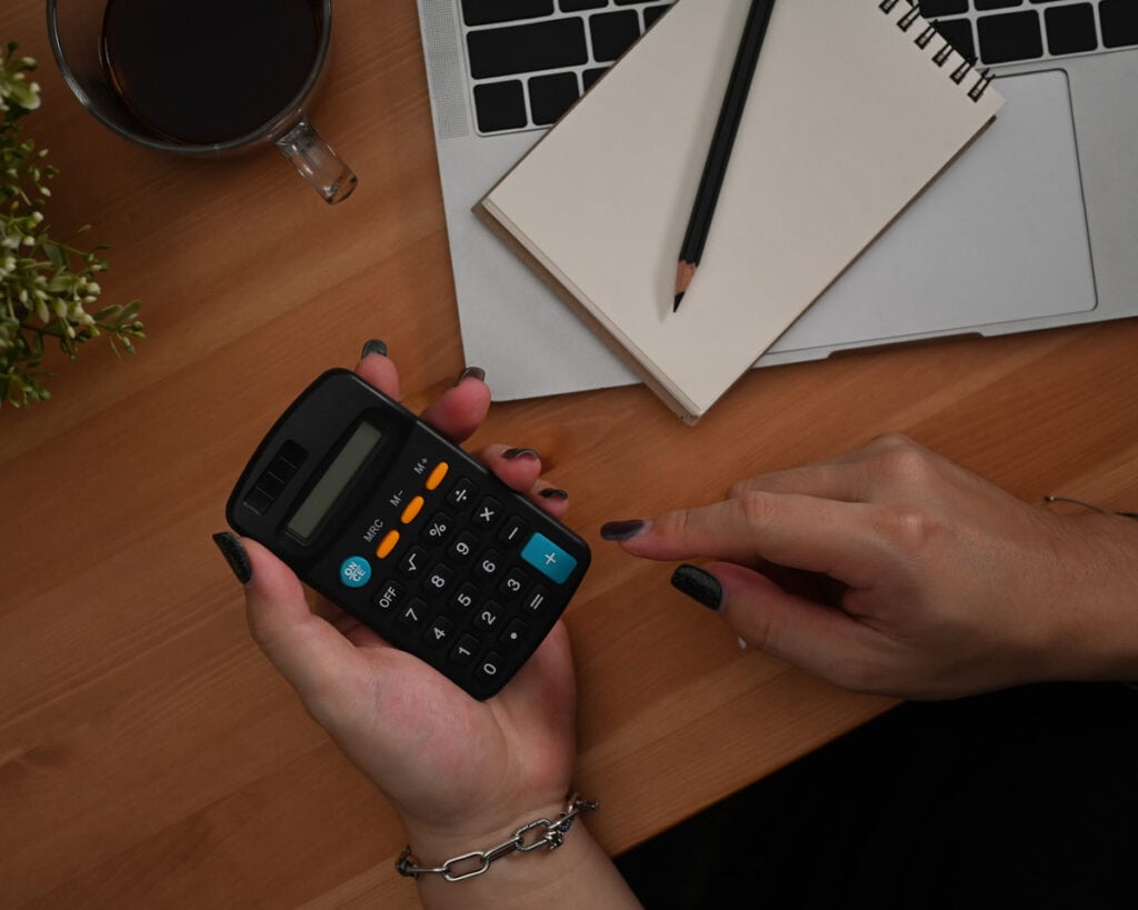 Person holding a calculator at a wooden desk with a laptop, notebook, pencil, plant, and cup of coffee.