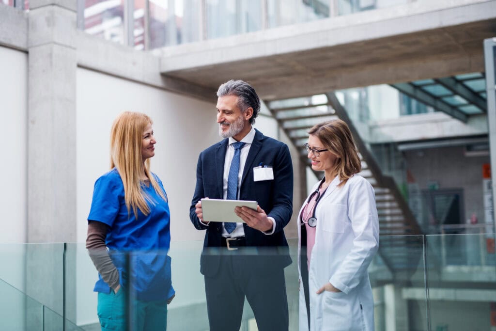 A group of three professionals in conversation, including a man in a suit holding a tablet, a woman in a blue nurse uniform, and another woman in a white doctor's coat with a stethoscope, stand in a modern building with glass and concrete elements.