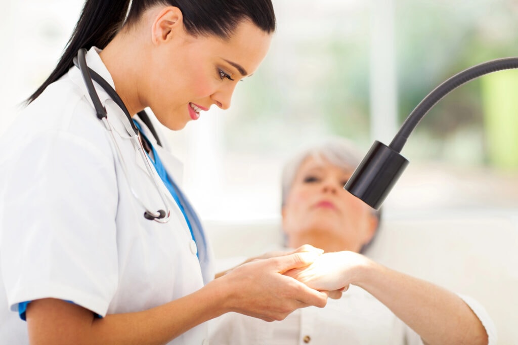 A dermatologist in a white coat examines the hand of an older woman under a bright lamp, utilizing hospital automation software to streamline care while the patient looks on in a modern medical office setting.