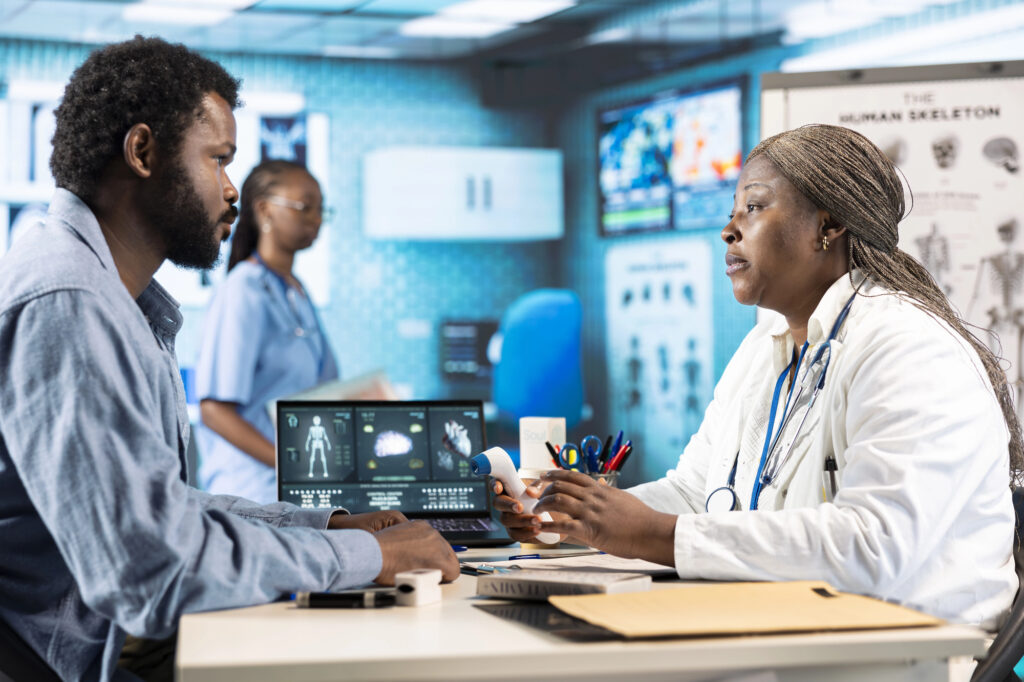 A doctor in a white coat consults with a male patient at a desk in a modern medical office, where physician contracts with hospitals and healthcare automation help streamline care. Another healthcare worker stands nearby, with charts and a skeleton poster visible.