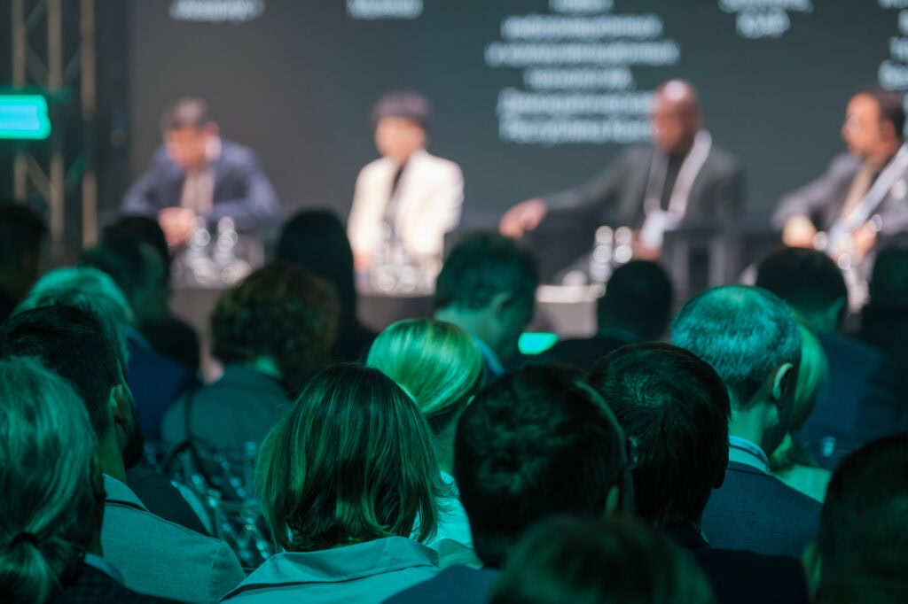An audience watches a panel of four people seated at a table on stage during a conference or discussion event, with the panelists and background slightly out of focus, as they explore topics like hospital automation.