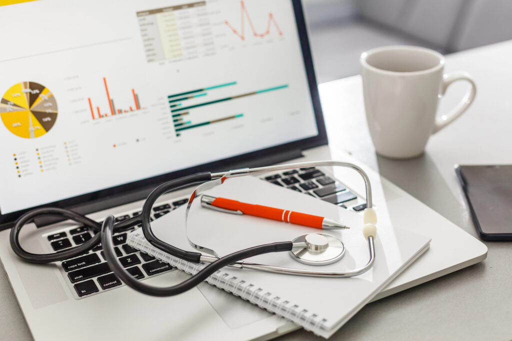 A stethoscope, notepad, and pen rest on a laptop keyboard displaying charts and graphs from a hospital automation system. A white coffee mug and smartphone sit nearby, all arranged neatly on a light-colored desk.