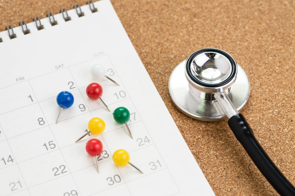 A stethoscope lies on a corkboard next to a calendar with colorful push pins marking key dates, suggesting health-related reminders managed efficiently with hospital automation software.