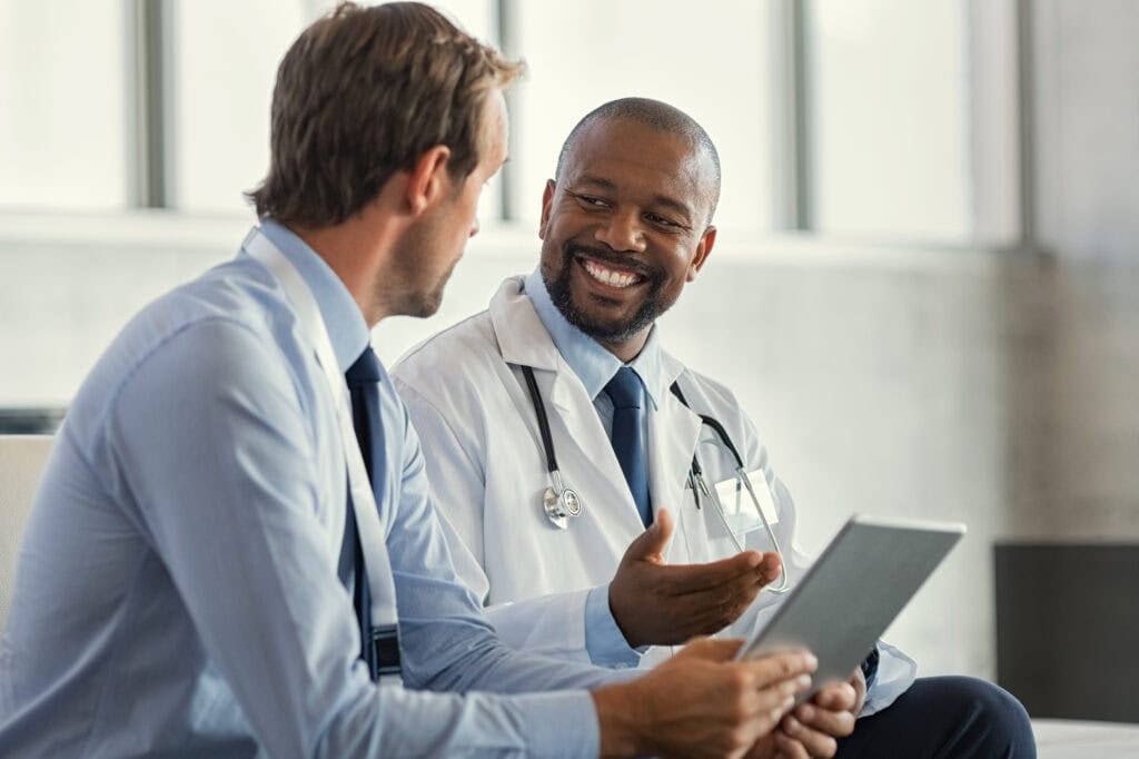 A smiling doctor in a white coat discusses healthcare compliance with a colleague in a suit, both holding digital tablets, in a bright, modern office setting.