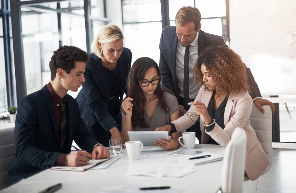 Five business professionals gathered around a table in a modern office, discussing physician contracts with hospitals and looking at a tablet, with papers, notebooks, and coffee cups on the table.