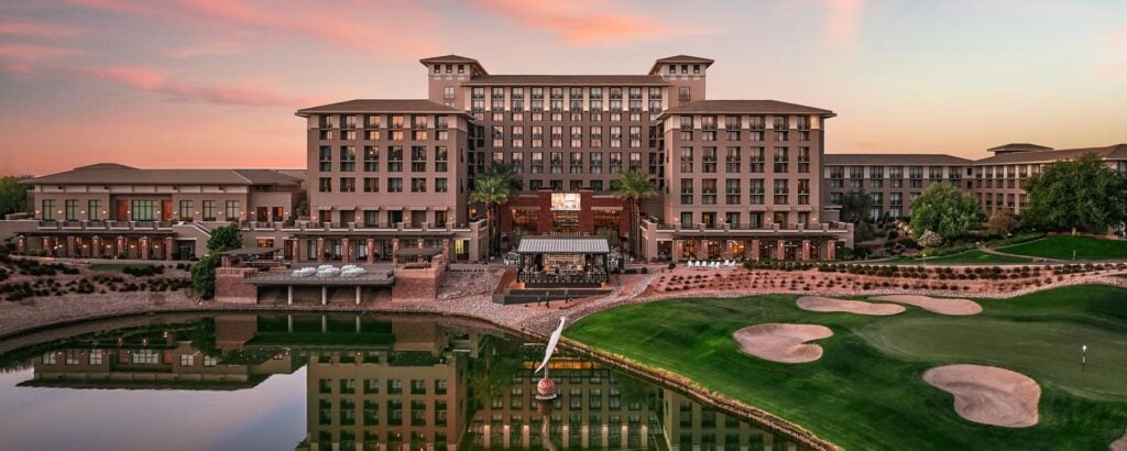 A large luxury hotel with multiple stories overlooks a golf course with sand traps and a small lake at sunset; pink clouds fill the sky.