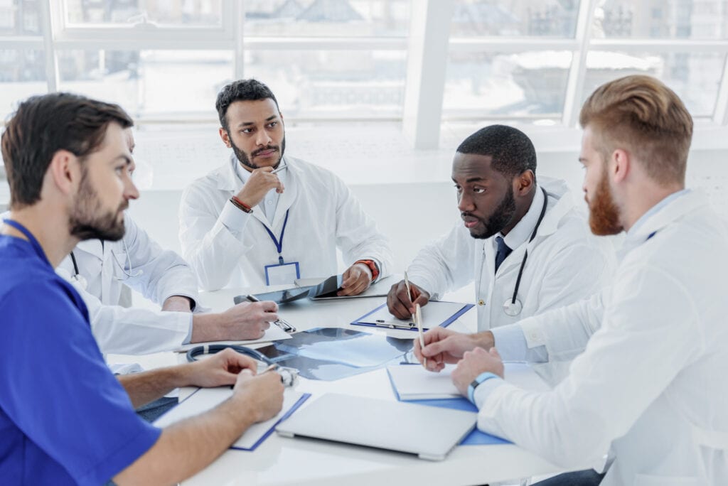 Five doctors in white coats sit around a table, discussing medical images and documents, as well as physician contracts with hospitals, in a bright, modern office with large windows.