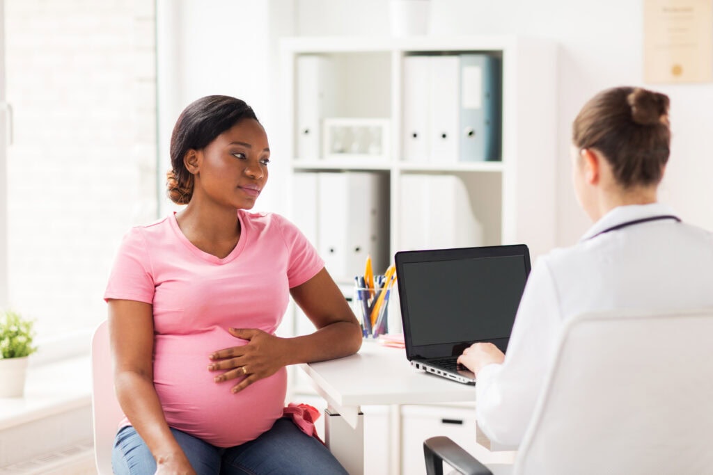 A pregnant woman in a pink shirt sits across from a doctor at a desk in a medical office, resting her hand on her belly while the doctor, discussing physician contracts with hospitals, types on a laptop.