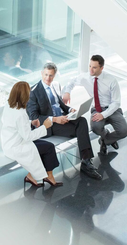 A female doctor in a lab coat in a meeting with two men in suits, one holding a laptop.