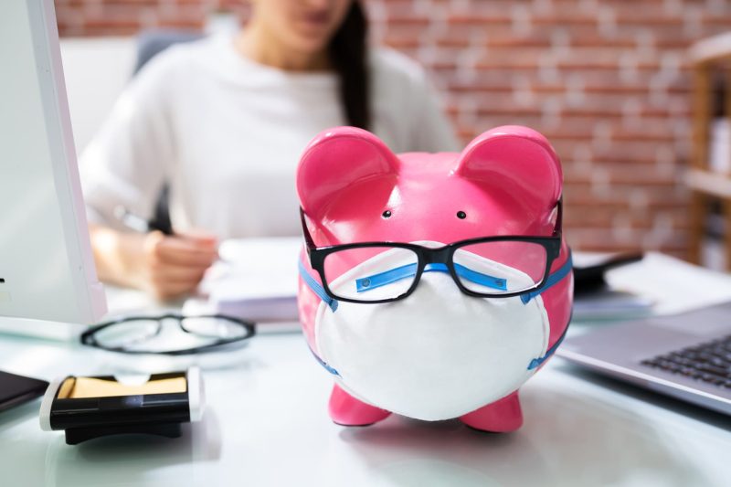 A piggy bank wearing a mask and glasses (signifying COVID-19) with a medical professional in the background